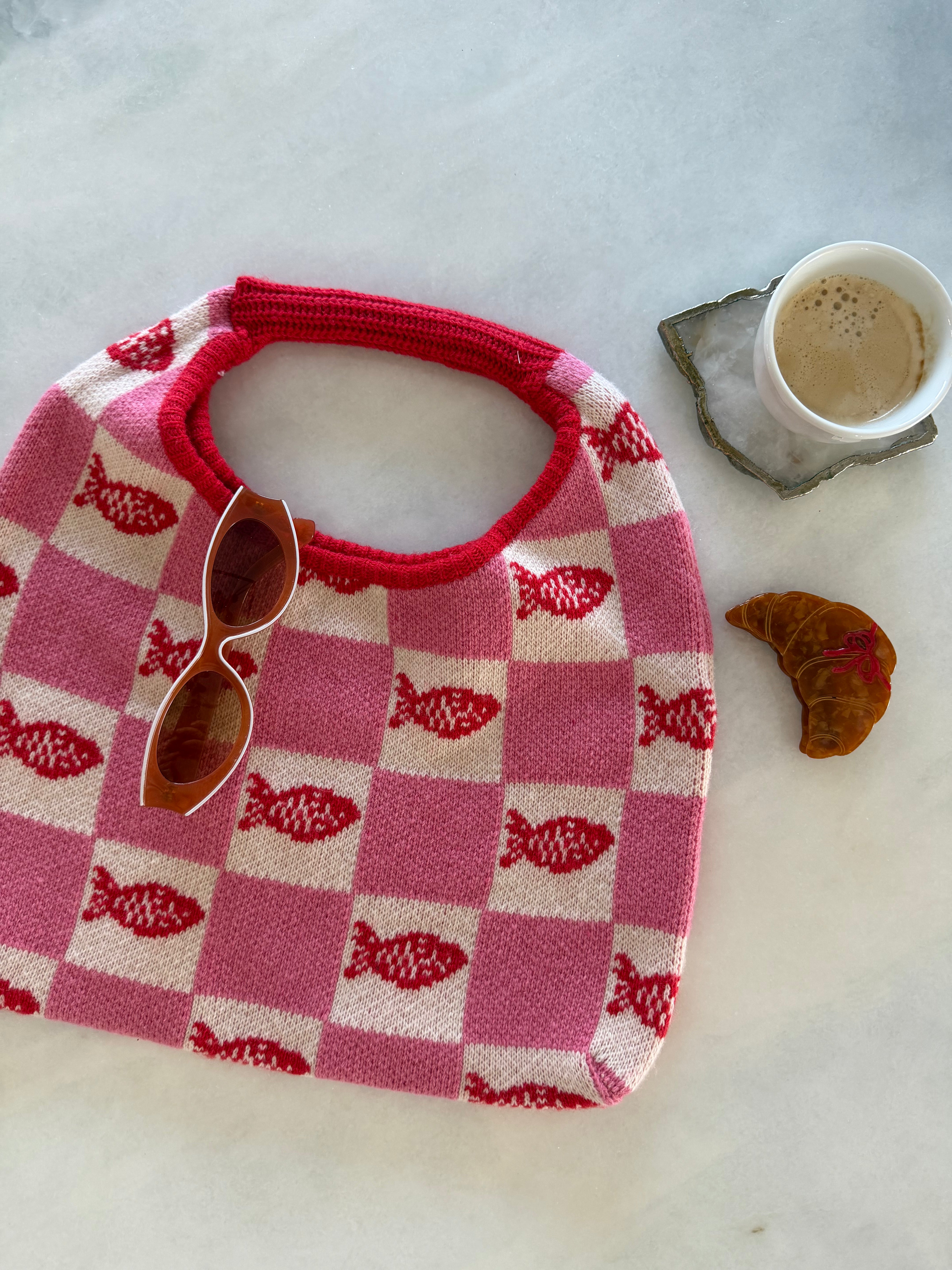 Pink and white checkered bib with red border on a marble surface, accompanied by sunglasses and a small cup. FRNCH Tulum Bag
