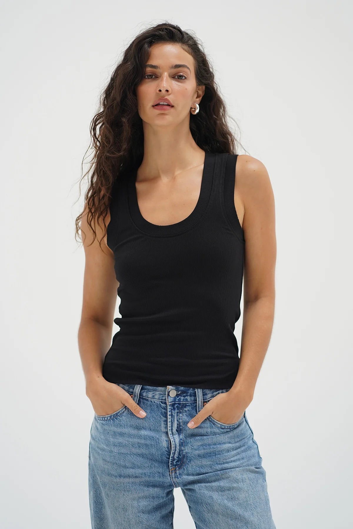 Woman wearing a black tank top and blue jeans against a white background. Woman wearing a white tank top and blue jeans against a plain background. LNA Double Layer Ribbed Scoop Tank at Coco Kimono Boutique