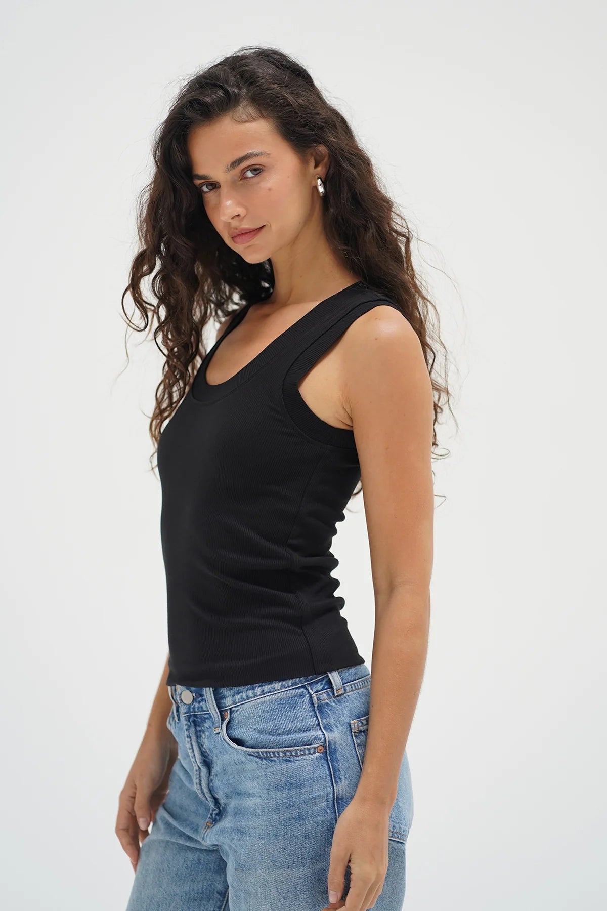 Woman wearing a black tank top and blue jeans against a white background. Woman wearing a white tank top and blue jeans against a plain background. LNA Double Layer Ribbed Scoop Tank at Coco Kimono Boutique